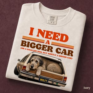 May include: An ivory t-shirt featuring the orange and brown text I NEED A BIGGER CAR (OR A SMALLER DOG, BUT MOSTLY A BIGGER CAR) above a graphic of a wood-panelled station wagon with a shaggy dog resting in the back and a license plate reading WOLFY.