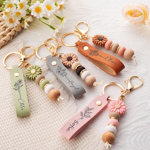 May include: Four personalised keychains featuring leather-like straps in sage green, grey, tan, and light pink. Each strap has a gold-tone metal ring, a floral line art engraving with a name, and a matching flower charm with wooden and silicone beads.