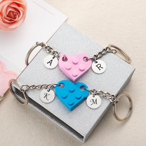 Peut inclure: Deux porte-clés en forme de cœur, un rose et un bleu, faits de blocs de construction. Chaque porte-clés a une chaîne argentée et un charme rond avec une lettre. Le cœur rose a des charmes "A" et "R", et le cœur bleu a des charmes "K" et "M".