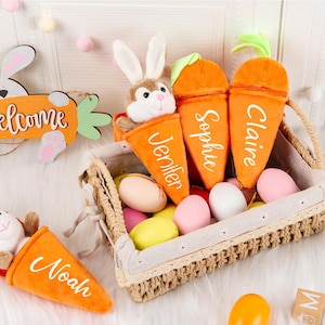 Può includere: Una composizione a tema pasquale con coniglietti di peluche in contenitori arancioni a forma di carota, ognuno con un nome. Un cestino intrecciato contiene uova colorate. È presente anche un cartello di legno con un coniglio e la parola "welcome".