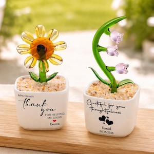 May include: Two glass flower figurines in white square pots. One features a yellow sunflower with the text 'Mrs Oswin thank you for helping me grow Emma'. The other features a green stem with purple bell-shaped flowers and the text 'Grateful for your thoughtfulness Daniel 06.15.2026'.