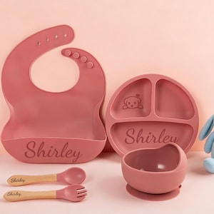 Könnte beinhalten: Ein Baby-Ess-Set in Staubrosa, bestehend aus Lätzchen, geteiltem Teller, Schüssel, Löffel und Gabel. Der Name "Shirley" ist eingraviert. Ein blaues Stoffkaninchen und eine kleine Figur sind ebenfalls im Bild zu sehen.