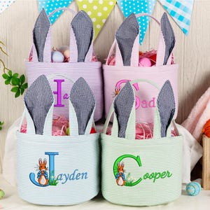 Peut inclure: Quatre paniers de Pâques avec des oreilles de lapin, chacun avec une couleur et une initiale différentes. Les paniers sont remplis d'herbe et d'œufs de Pâques. Un panier porte le nom "Jayden" brodé, et un autre le nom "Cooper". Un lapin en peluche et des carottes sont en arrière-plan.