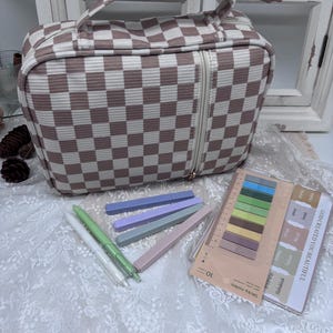 May include: A brown and white checkered cosmetic bag with a zipper closure and a handle. Beside the bag are several pens, sticky notes, and highlighters. The sticky notes have the text "GOD CREATED YOU BEAUTIFUL".