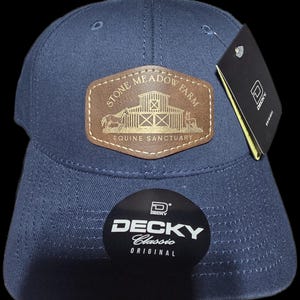 May include: A navy blue baseball cap with a curved brim. The cap features a brown leather patch with the text "STONE MEADOW FARM EQUINE SANCTUARY" and a barn graphic. A black tag with the "DECKY" logo is attached.