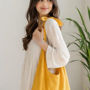 May include: A bright yellow tote bag with a rounded shape and a single pocket. The bag is carried over the shoulder with a matching yellow bow detail. The background is a neutral color with a green plant.