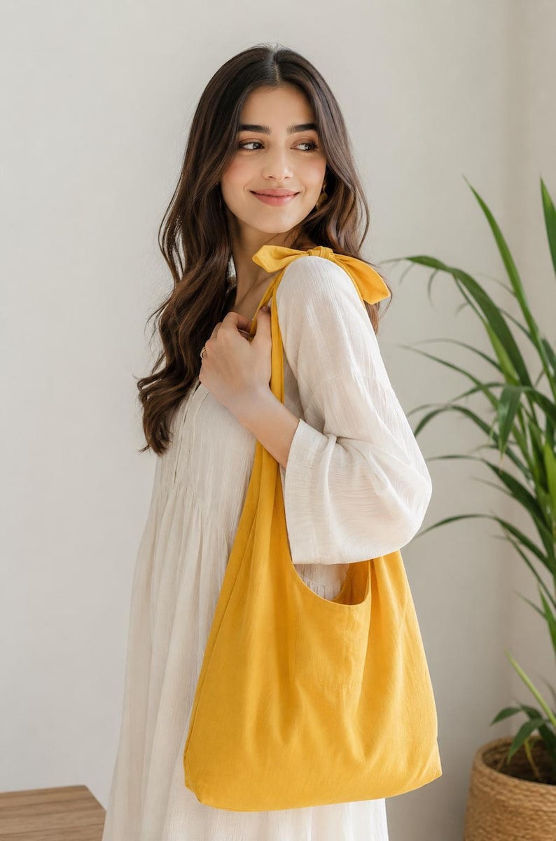 May include: A bright yellow tote bag with a rounded shape and a single pocket. The bag is carried over the shoulder with a matching yellow bow detail. The background is a neutral color with a green plant.