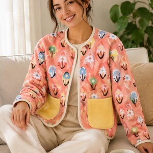 May include: A coral-colored jacket with a floral pattern in blue, yellow, and white. The jacket has a cream-colored trim, two yellow pockets, and is paired with cream-colored pants. A cup of coffee and cookies are on a table.