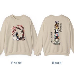 May include: Beige sweatshirt featuring a front graphic of characters under a flowering tree. The back displays a stacked character design with Japanese text and "STUDIO GHIBLI" in white. A cozy, casual garment.