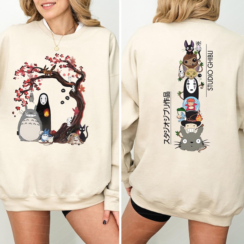 May include: Beige sweatshirt featuring a colorful Studio Ghibli design. The front displays characters under a flowering tree. The back showcases a stacked arrangement of characters with "STUDIO GHIBLI" and Japanese text.