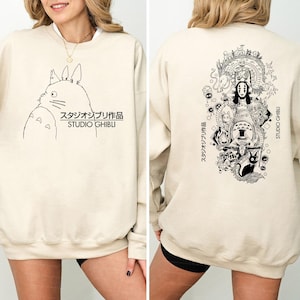 May include: Beige sweatshirt with a black and white Studio Ghibli character illustration on the front. The text "スタジオジブリ作品 STUDIO GHIBLI" is printed below the character. The back of the sweatshirt features a larger illustration of various Studio Ghibli characters.