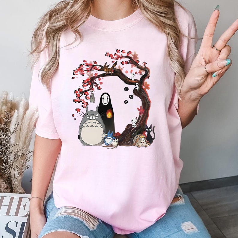 May include: A light pink t-shirt featuring a colorful graphic of a tree with red blossoms and various animated characters. The characters include a large gray creature, a black figure, and several smaller creatures. The design is centered on the shirt.