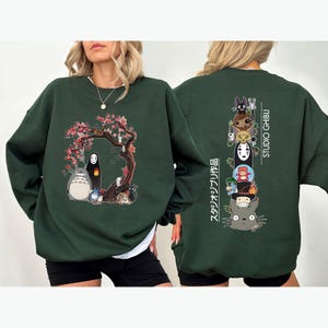 May include: Dark green sweatshirt featuring a colorful graphic of characters from Studio Ghibli films. The front design includes Totoro, No-Face, and other figures under a flowering tree. The back displays a vertical stack of characters with "STUDIO GHIBLI" and Japanese text.
