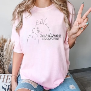 May include: A pale pink t-shirt with a black illustration of two cartoon characters and the text "スタジオジブリ作品 STUDIO GHIBLI". The shirt has short sleeves.