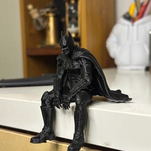 以下が含まれることがあります： マントを羽織ったスーパーヒーローの黒い3Dプリントフィギュア。前かがみの姿勢で座っており、詳細なマスク、胸部と四肢のテクスチャ加工されたアーマー、そして表面の端から垂れ下がる長いマントが特徴です。