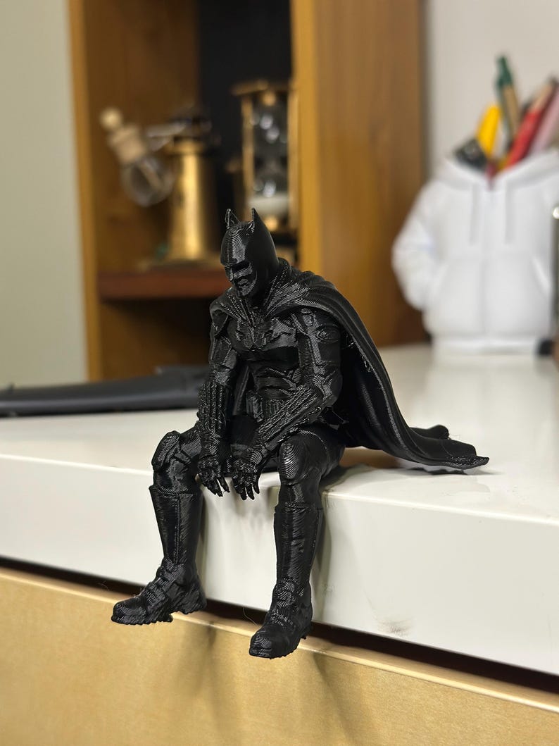 以下が含まれることがあります： マントを羽織ったスーパーヒーローの黒い3Dプリントフィギュア。前かがみの姿勢で座っており、詳細なマスク、胸部と四肢のテクスチャ加工されたアーマー、そして表面の端から垂れ下がる長いマントが特徴です。