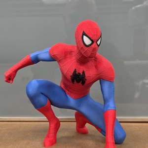 May include: A red, blue, and black Spider-Man figurine in a crouching pose. The figurine has a red mask with white eye lenses, a red torso, blue arms and legs, and red gloves and boots. A black spider emblem is on the chest.