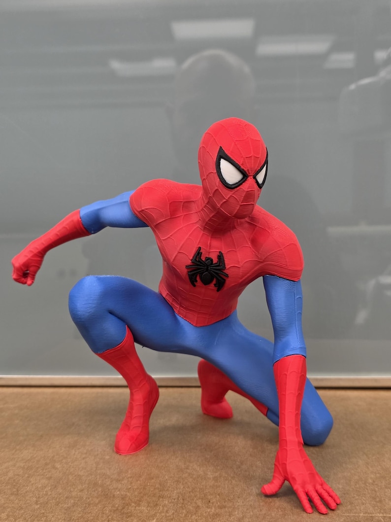 May include: A red, blue, and black Spider-Man figurine in a crouching pose. The figurine has a red mask with white eye lenses, a red torso, blue arms and legs, and red gloves and boots. A black spider emblem is on the chest.
