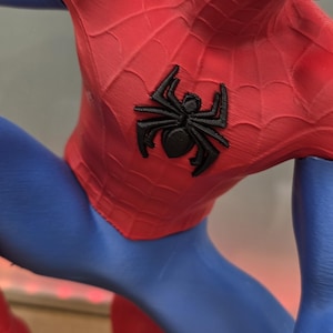 May include: A close-up of a red and blue Spider-Man costume. The red portion features a web pattern and a black spider emblem on the chest. The blue sections are smooth and form-fitting.