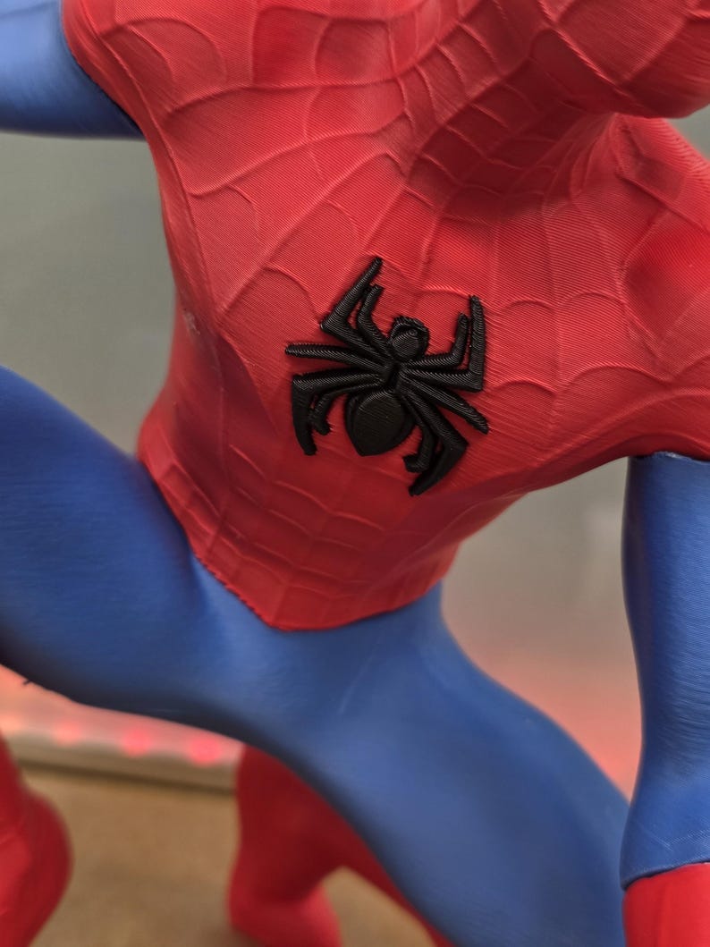 May include: A close-up of a red and blue Spider-Man costume. The red portion features a web pattern and a black spider emblem on the chest. The blue sections are smooth and form-fitting.