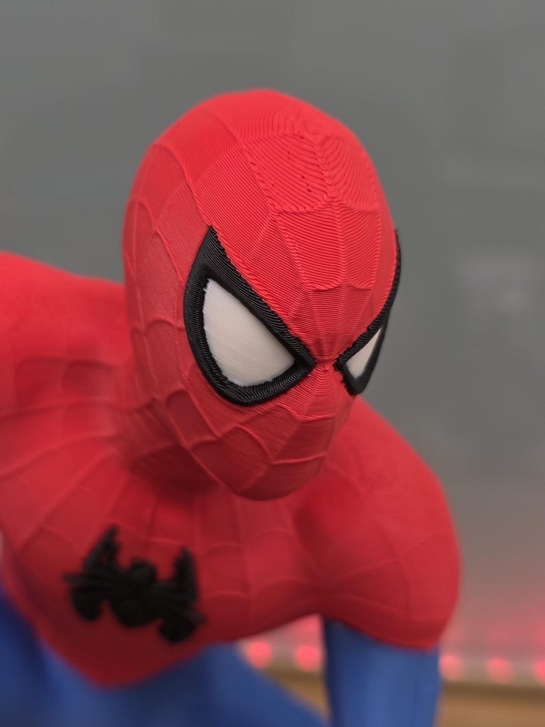 May include: A red and blue Spider-Man figurine. The figure has a red mask with white eyes outlined in black. The costume is red with a black spider emblem on the chest and blue on the lower body.
