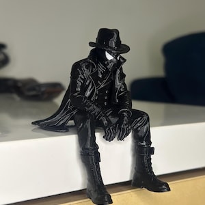 May include: A black 3D printed figurine of a character wearing a fedora, a long trench coat, and lace-up boots. The figure is posed in a seated position with legs dangling, featuring a mask with white eye lenses and a textured, monochromatic finish.