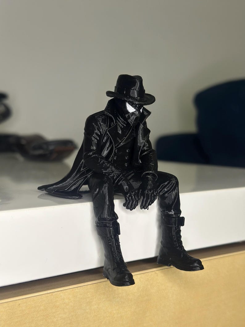 May include: A black 3D printed figurine of a character wearing a fedora, a long trench coat, and lace-up boots. The figure is posed in a seated position with legs dangling, featuring a mask with white eye lenses and a textured, monochromatic finish.
