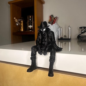 May include: A black 3D printed figurine of a character wearing a trench coat, a fedora hat, and boots, sitting with legs dangling. The figure features a textured black finish, a high collar, and distinct white eye lenses on the mask, capturing a noir-inspired aesthetic in a seated pose.