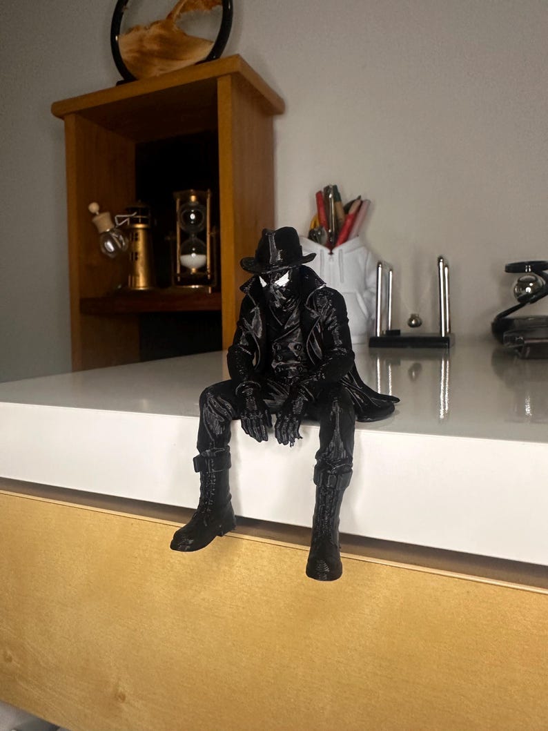 May include: A black 3D printed figurine of a character wearing a trench coat, a fedora hat, and boots, sitting with legs dangling. The figure features a textured black finish, a high collar, and distinct white eye lenses on the mask, capturing a noir-inspired aesthetic in a seated pose.