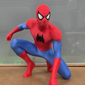 May include: A red, blue, and black Spider-Man figurine in a crouching pose. The figure has a red mask with white eye lenses, a red torso with a black spider emblem, blue legs, and red gloves and boots. The figure is a collectible.