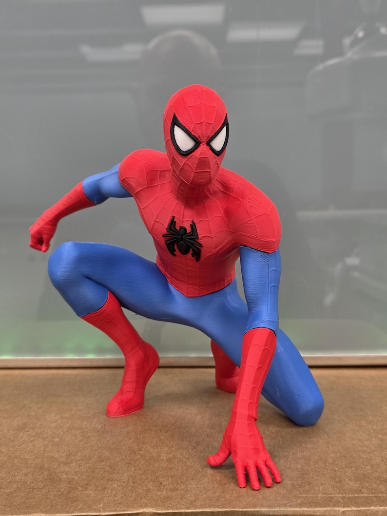 May include: A red, blue, and black Spider-Man figurine in a crouching pose. The figure has a red mask with white eye lenses, a red torso with a black spider emblem, blue legs, and red gloves and boots. The figure is a collectible.