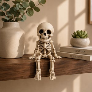 May include: A beige skeleton figurine with a large, rounded skull and detailed ribcage, sitting with legs dangling over the edge. The figure has hollow eye sockets and a visible jawline, crafted in a neutral tone to resemble bone structure.