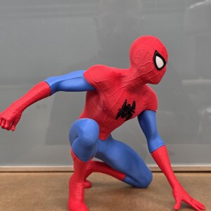 May include: A red and blue Spider-Man figurine in a crouching pose. The figure has a red mask with white eye lenses, red arms, and red boots. The torso and legs are blue, with a black spider emblem on the chest.