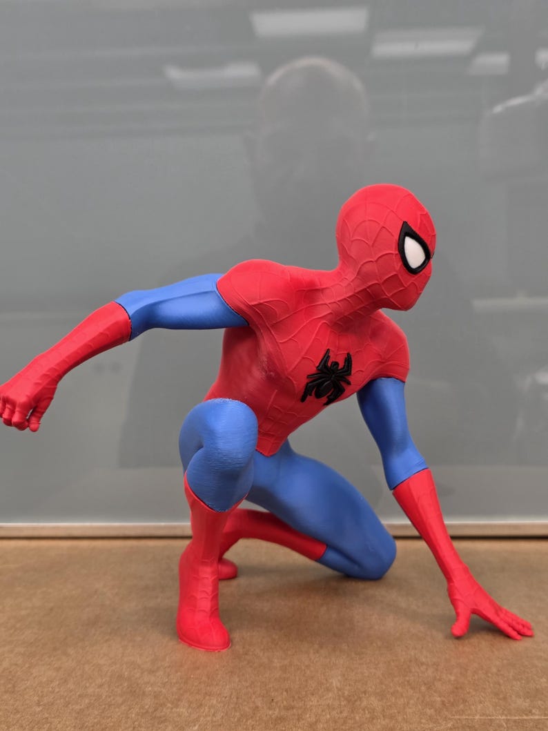 May include: A red and blue Spider-Man figurine in a crouching pose. The figure has a red mask with white eye lenses, red arms, and red boots. The torso and legs are blue, with a black spider emblem on the chest.