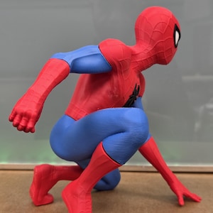 May include: A red, blue, and black Spider-Man figurine in a crouching pose. The figure is wearing a red and blue suit with a black spider emblem on the chest and a red mask with white eye lenses. The figure is in a dynamic, action-oriented stance.