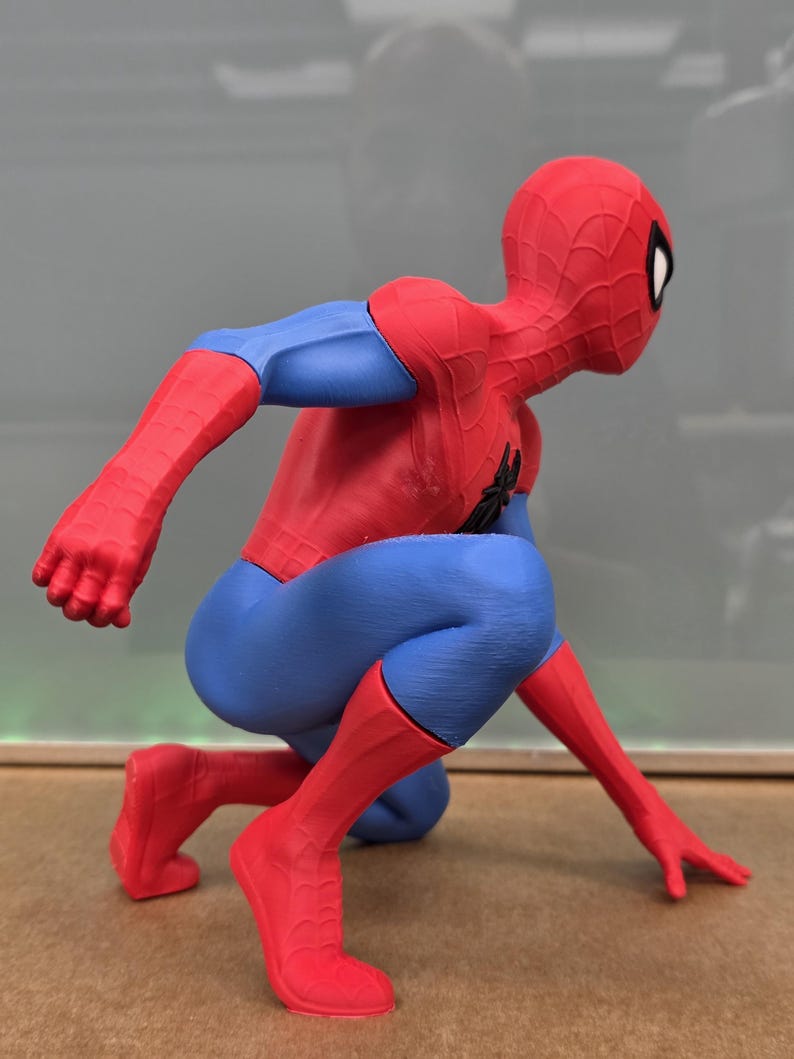May include: A red, blue, and black Spider-Man figurine in a crouching pose. The figure is wearing a red and blue suit with a black spider emblem on the chest and a red mask with white eye lenses. The figure is in a dynamic, action-oriented stance.