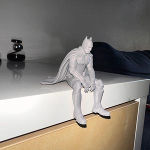 以下が含まれることがあります： マントを羽織ったスーパーヒーローのライトグレーの3Dプリントフィギュア。脚を縁からぶら下げて座っている。尖ったマスク、腕と脚に装甲板が付いた質感のあるスーツ、背中に垂れ下がった長いマントが特徴である。