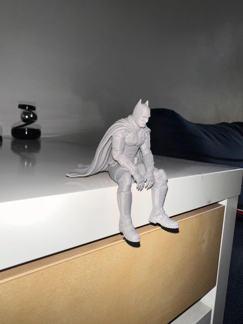 以下が含まれることがあります： マントを羽織ったスーパーヒーローのライトグレーの3Dプリントフィギュア。脚を縁からぶら下げて座っている。尖ったマスク、腕と脚に装甲板が付いた質感のあるスーツ、背中に垂れ下がった長いマントが特徴である。