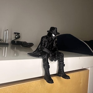 May include: A black figurine of a character wearing a trench coat, a fedora hat, and boots, depicted in a seated position with legs dangling. The figure features a mask with white eye lenses and is rendered in a solid, monochromatic black finish.