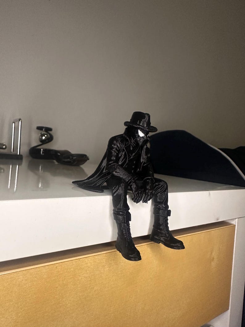 May include: A black figurine of a character wearing a trench coat, a fedora hat, and boots, depicted in a seated position with legs dangling. The figure features a mask with white eye lenses and is rendered in a solid, monochromatic black finish.