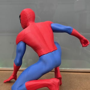 May include: A red, blue, and black Spider-Man action figure in a crouching pose. The figure is wearing a red and blue suit with a spiderweb pattern on the head. The figure is kneeling on a brown surface.