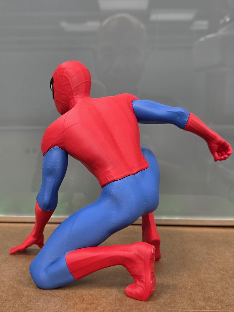 May include: A red, blue, and black Spider-Man action figure in a crouching pose. The figure is wearing a red and blue suit with a spiderweb pattern on the head. The figure is kneeling on a brown surface.