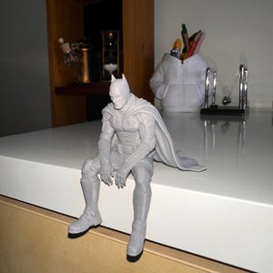 以下が含まれることがあります： マントを羽織ったスーパーヒーローのライトグレーの3Dプリントフィギュア。縁に腰掛け、足をぶら下げた姿勢をとっています。フィギュアには詳細なマスク、質感のあるガントレット、そして座った姿勢の後ろに伸びる流れるようなマントが表現されています。