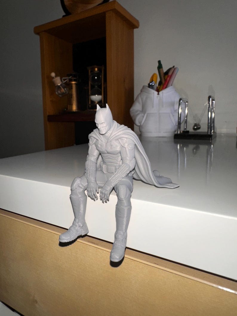 以下が含まれることがあります： マントを羽織ったスーパーヒーローのライトグレーの3Dプリントフィギュア。縁に腰掛け、足をぶら下げた姿勢をとっています。フィギュアには詳細なマスク、質感のあるガントレット、そして座った姿勢の後ろに伸びる流れるようなマントが表現されています。