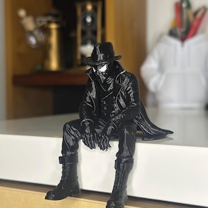May include: A black figurine of a character wearing a fedora, a long trench coat, and tall boots, sitting with legs dangling. The figure has white eye lenses and is sculpted with detailed textures on the clothing, including buttons and fabric folds.