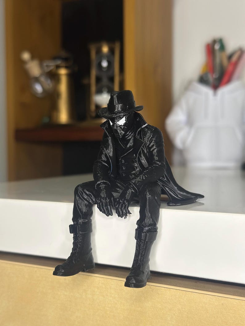 May include: A black figurine of a character wearing a fedora, a long trench coat, and tall boots, sitting with legs dangling. The figure has white eye lenses and is sculpted with detailed textures on the clothing, including buttons and fabric folds.