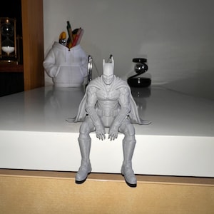 以下が含まれることがあります： 縁から足をぶら下げて座っている、ライトグレーの3Dプリントされたバットマンのフィギュア。キャラクターは、肩と背中にマントを羽織り、尖ったカウルとユーティリティベルトを備えた、質感のある詳細なスーツを着用しています。