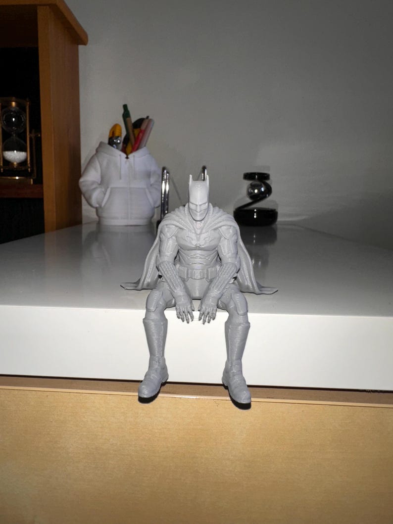以下が含まれることがあります： 縁から足をぶら下げて座っている、ライトグレーの3Dプリントされたバットマンのフィギュア。キャラクターは、肩と背中にマントを羽織り、尖ったカウルとユーティリティベルトを備えた、質感のある詳細なスーツを着用しています。