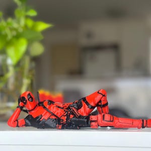 May include: A red and black Deadpool figurine in a reclining pose, resting on one elbow with legs extended. The character wears a full-body suit with black harness details, a utility belt, and a mask with white eye patches, rendered in a smooth finish.