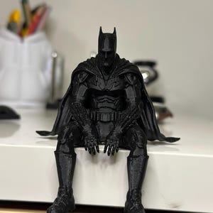 以下が含まれることがあります： 肩にマントを羽織り、座った姿勢をとるバットマンの黒い3Dプリントフィギュア。フィギュア全体に詳細なアーマープレート、尖ったカウル、質感のある表面が施されており、キャラクターの全身が精巧に再現されています。